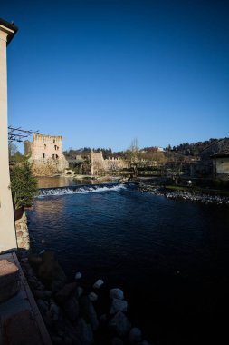 İtalya 'nın kırsal kesimindeki bir köyden geçen nehir, kıyılarında evler olan ve üzerinde kaleler ve eski metalik bir yol köprüsü bulunan bir sınır duvarının kalıntıları olan arka planda.