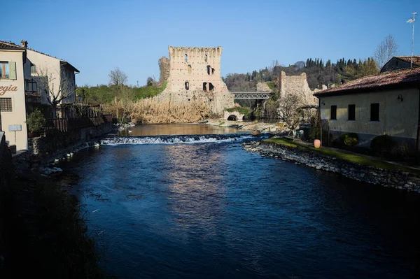 İtalya 'nın kırsal kesimindeki bir köyden geçen nehir, kıyılarında evler olan ve üzerinde kaleler ve eski metalik bir yol köprüsü bulunan bir sınır duvarının kalıntıları olan arka planda.