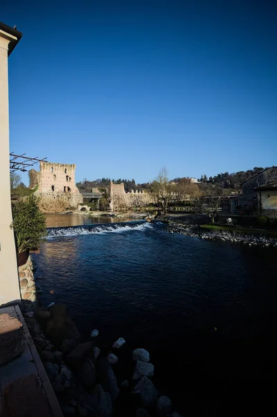 İtalya 'nın kırsal kesimindeki bir köyden geçen nehir, kıyılarında evler olan ve üzerinde kaleler ve eski metalik bir yol köprüsü bulunan bir sınır duvarının kalıntıları olan arka planda.