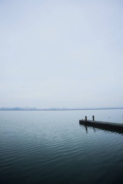Bir gölde uzanan ahşap demirleme direkleriyle rıhtım, bulutlu ve sisli bir günde, karşı kıyıda, İtalya kırsalında