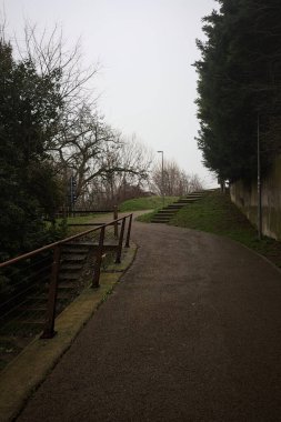 Bir yamaçta bisiklet yolu, ağaçlarla ve sınır duvarıyla çevrili, yolun kenarında küçük beton bir merdiven, bir İtalyan kasabasında bulutlu bir günde