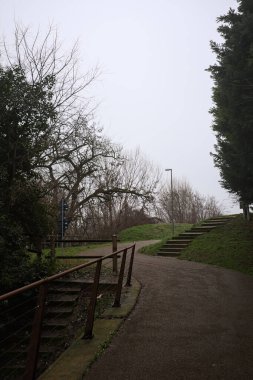 Bir yamaçta bisiklet yolu, ağaçlarla ve sınır duvarıyla çevrili, yolun kenarında küçük beton bir merdiven, bir İtalyan kasabasında bulutlu bir günde