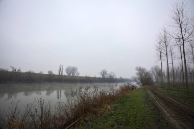 Bulutlu ve sisli bir günde, İtalya 'nın kırsalında, nehrin kıyısındaki bir parkta, karşı kıyıda ağaçların olduğu bir patika.