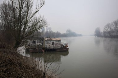 Çamurlu bir koruluğun altındaki nehir kıyısı, demirli bir yüzen ev, suya yansıyan ağaçlar, ve İtalya 'nın arka planındaki zıt kıyılar, bulutlu ve sisli bir günde görülür.