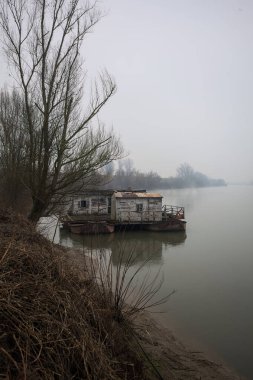 Çamurlu bir koruluğun altındaki nehir kıyısı, demirli bir yüzen ev, suya yansıyan ağaçlar, ve İtalya 'nın arka planındaki zıt kıyılar, bulutlu ve sisli bir günde görülür.