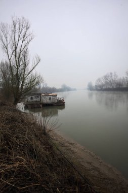 Çamurlu bir koruluğun altındaki nehir kıyısı, demirli bir yüzen ev, suya yansıyan ağaçlar, ve İtalya 'nın arka planındaki zıt kıyılar, bulutlu ve sisli bir günde görülür.