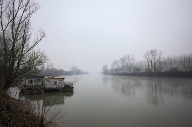 Çamurlu bir koruluğun altındaki nehir kıyısı, demirli bir yüzen ev, suya yansıyan ağaçlar, ve İtalya 'nın arka planındaki zıt kıyılar, bulutlu ve sisli bir günde görülür.