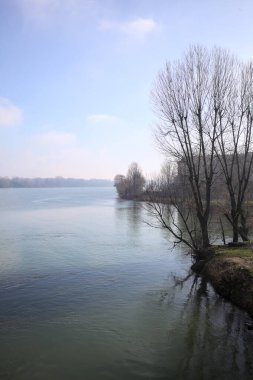 Park, çıplak ağaçlarla, güneşli bir günde göl kıyısında, gökler suya dökülür ve karşı kıyılar uzak mesafede olur.