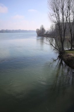 Park, çıplak ağaçlarla, güneşli bir günde göl kıyısında, gökler suya dökülür ve karşı kıyılar uzak mesafede olur.