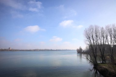 Park, çıplak ağaçlarla, güneşli bir günde göl kıyısında, gökler suya dökülür ve karşı kıyılar uzak mesafede olur.