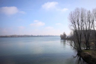 Park, çıplak ağaçlarla, güneşli bir günde göl kıyısında, gökler suya dökülür ve karşı kıyılar uzak mesafede olur.
