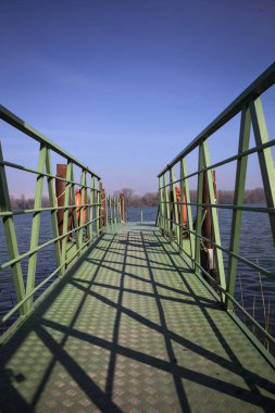 Bir rıhtımın Jetty 'si, bir gölün üzerine uzanan, ucunda demirleme direkleriyle, ve uzakta karşı kıyıda, güneşli bir kış gününde görülen