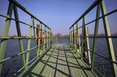 Bir rıhtımın Jetty 'si, bir gölün üzerine uzanan, ucunda demirleme direkleriyle, ve uzakta karşı kıyıda, güneşli bir kış gününde görülen