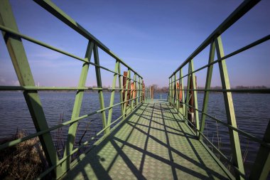 Bir rıhtımın Jetty 'si, bir gölün üzerine uzanan, ucunda demirleme direkleriyle, ve uzakta karşı kıyıda, güneşli bir kış gününde görülen