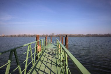 Bir rıhtımın Jetty 'si, bir gölün üzerine uzanan, ucunda demirleme direkleriyle, ve uzakta karşı kıyıda, güneşli bir kış gününde görülen