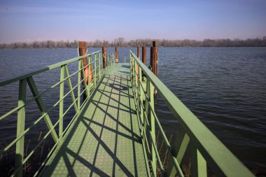 Bir rıhtımın Jetty 'si, bir gölün üzerine uzanan, ucunda demirleme direkleriyle, ve uzakta karşı kıyıda, güneşli bir kış gününde görülen