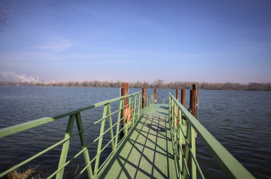 Bir rıhtımın Jetty 'si, bir gölün üzerine uzanan, ucunda demirleme direkleriyle, ve uzakta karşı kıyıda, güneşli bir kış gününde görülen