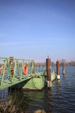 Bir rıhtımın Jetty 'si, bir gölün üzerine uzanan, ucunda demirleme direkleriyle, ve uzakta karşı kıyıda, güneşli bir kış gününde görülen