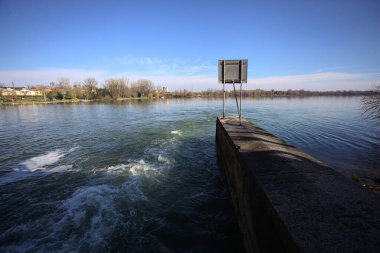 Güneşli bir günde, bir su kanalının sonundan, akan suyla, bir gölün karşı kıyısında, arkasında işaret olan beton bir duvar.