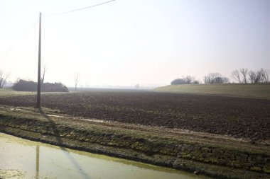 Pürüzlü tarlalar, ufukta ağaçlar, güneşli bir günde setle çevrili, su dolu bir siper ve ahşap sütunlar üzerinde elektrik kabloları ile çevrili.