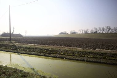 Pürüzlü tarlalar, ufukta ağaçlar, güneşli bir günde setle çevrili, su dolu bir siper ve ahşap sütunlar üzerinde elektrik kabloları ile çevrili.