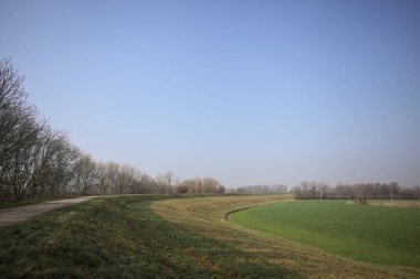 Bir tarafta çıplak ağaçlarla çevrili çamurlu bir set ve İtalya kırsalında güneşli bir günde yamaçlarda uzanan eğimli bir arazi.