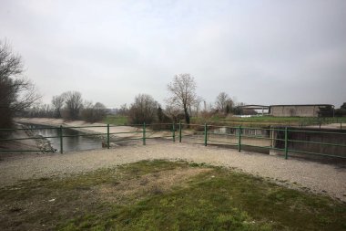 Teras, ağaçlar arasındaki sıra sıra saptırıcı bir kanal üzerinde demiryolu ile çevrili ve İtalya kırsalında bulutlu bir günde balyalarla kaplı bir ahır var.