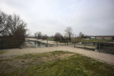 Teras, ağaçlar arasındaki sıra sıra saptırıcı bir kanal üzerinde demiryolu ile çevrili ve İtalya kırsalında bulutlu bir günde balyalarla kaplı bir ahır var.