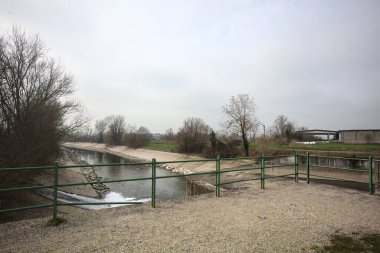 Teras, ağaçlar arasındaki sıra sıra saptırıcı bir kanal üzerinde demiryolu ile çevrili ve İtalya kırsalında bulutlu bir günde balyalarla kaplı bir ahır var.