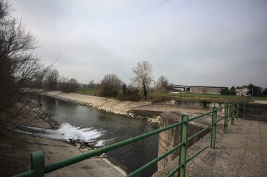 Teras, ağaçlar arasındaki sıra sıra saptırıcı bir kanal üzerinde demiryolu ile çevrili ve İtalya kırsalında bulutlu bir günde balyalarla kaplı bir ahır var.