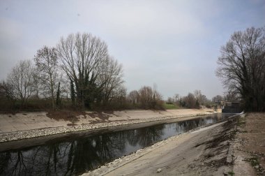İtalya kırsalında bulutlu bir günde, kıyıları çevreleyen çıplak ağaçlarla, gökyüzü ile birlikte suya yansıyan saptırıcı kanal.