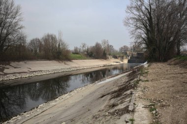 İtalya kırsalında bulutlu bir günde, kıyıları çevreleyen çıplak ağaçlarla, gökyüzü ile birlikte suya yansıyan saptırıcı kanal.