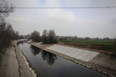 İtalya kırsalında bulutlu bir günde, kıyıları çevreleyen çıplak ağaçlarla, gökyüzü ile birlikte suya yansıyan saptırıcı kanal.