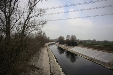 İtalya kırsalında bulutlu bir günde, kıyıları çevreleyen çıplak ağaçlarla, gökyüzü ile birlikte suya yansıyan saptırıcı kanal.