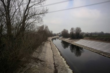 İtalya kırsalında bulutlu bir günde, kıyıları çevreleyen çıplak ağaçlarla, gökyüzü ile birlikte suya yansıyan saptırıcı kanal.