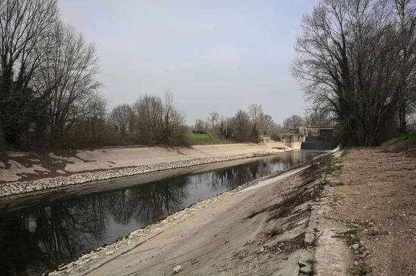 İtalya kırsalında bulutlu bir günde, kıyıları çevreleyen çıplak ağaçlarla, gökyüzü ile birlikte suya yansıyan saptırıcı kanal.