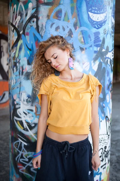 Beautiful hip teenage blonde woman standing by graffiti wall — Stock Photo, Image