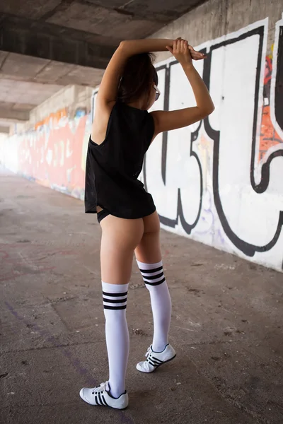 Hermosa mujer con gafas de sol negras y ropa deportiva negra bailando sexy frente a la pared de graffiti — Foto de Stock