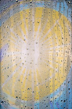 Su damlaları, soyut, parlak sarı-mavi bir zemin üzerinde şeffaf bir camın yüzeyinden aşağı akar..