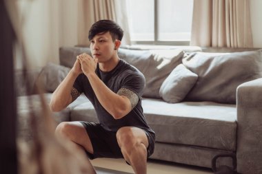 Spor giyimli formalı adam evde çömelme egzersizi yapıyor. Genç Asyalı adam evde antrenman yapıyor. Karantinada ev egzersizi.