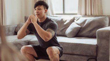 Spor giyimli formalı adam evde çömelme egzersizi yapıyor. Genç Asyalı adam evde antrenman yapıyor. Karantinada ev egzersizi.