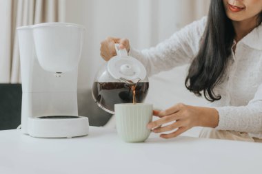 Genç mutlu kadın evde kahve makinesi kullanıyor. Asyalı kadın bir fincana kahve doldurup evde kahve yapıyor..