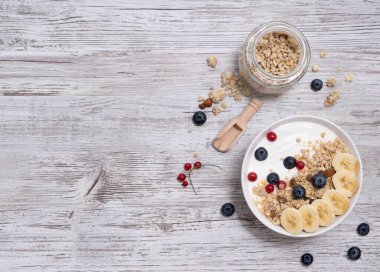 Tahta bir masada bir kase müsli, muz, yaban mersini ve kırmızı frenk üzümü. Düşüncelerle dolu bir kavanoz ve tahta bir kaşık.