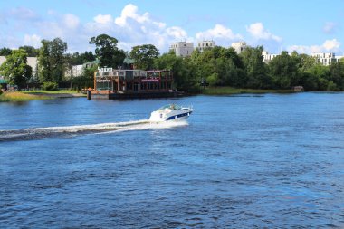 Nehirde yüzen bir restoranın arka planına, ağaçlara ve bulutlu mavi gökyüzüne karşı giden beyaz bir tekne. St. Petersburg