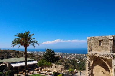 Bellapais Manastırı 'nın duvarının bir kısmı görünüyor. Aşağıda bir restoran ve bir palmiye ağacı var. Ufukta, Kyrenia şehri, deniz ve parlak mavi gökyüzü. Kyrenia. Kıbrıs.
