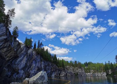 Kayalık sahilin altından bir manzara ve güneşli bir yaz gününde Ruskeala Dağ Parkı 'ndaki Mermer Kanyon' un zümrüt suyuna düşen büyük bir kaya..