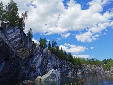 Kayalık sahilin altından bir manzara ve güneşli bir yaz gününde Ruskeala Dağ Parkı 'ndaki Mermer Kanyon' un zümrüt suyuna düşen büyük bir kaya..