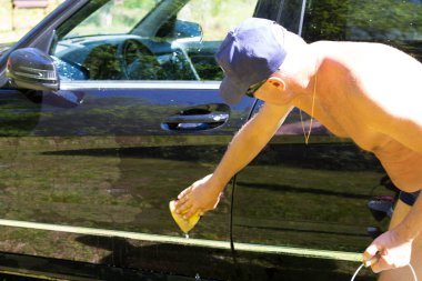 Yüzme şortlu beyaz bir adam, siyah tenli sahibinin arabasını manuel olarak süngerle temizler..