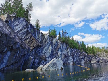 Kayalık sahilin altından bir manzara ve güneşli bir yaz gününde Ruskeala Dağ Parkı 'ndaki Mermer Kanyon' un zümrüt suyuna düşen büyük bir kaya..