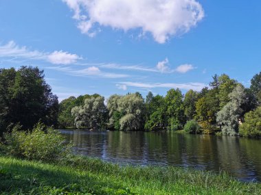 Gölün kıyısı, insanların kayıkla gezdiği yer, ağaçlarla çevrili, güneşli bir yaz gününde bulutlu mavi gökyüzünün arka planına karşı..